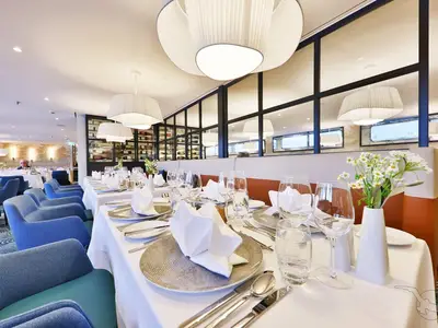Das elegante Restaurant der MS Amina mit festlich gedeckten Tischen und blauen Sesseln. Moderne Beleuchtung und ein Weinregal prägen das stilvolle Ambiente.