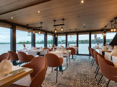 Elegantes Restaurant an Bord der MS Amina mit braunen Stühlen und weiß gedeckten Tischen. Panoramablick durch große Fenster auf einen Fluss und die Uferlandschaft.