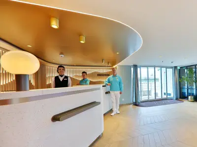 Die moderne Rezeption der MS Amina mit lächelnden Crew-Mitgliedern. Goldene Decke, ovales Lichtobjekt und große Fensterfront für Tageslicht.