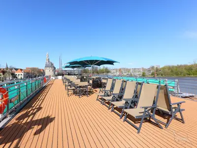 Das Sonnendeck der MS Amina an einem sonnigen Tag mit Liegestühlen, Tischen und blauen Sonnenschirmen. Im Hintergrund eine Stadt mit historischen Gebäuden und ein Fluss.