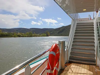 Blick vom Sonnendeck der MS Amina auf den glitzernden Rhein und bewaldete Berge. Eine Treppe führt zu einer oberen Ebene, ein Rettungsring hängt an der Reling.