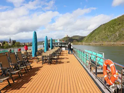 Das Sonnendeck der MS Amina zeigt Liegestühle und Tische. Mehrere Passagiere genießen die Rheinfahrt, mit Blick auf grüne Berge, ein Dorf und eine Burg.