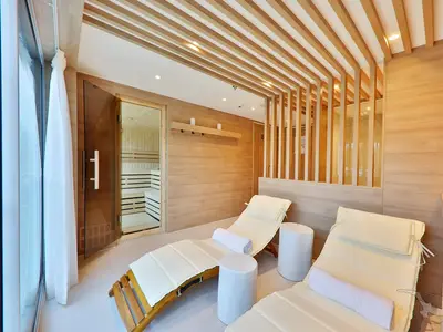 Ein heller, moderner Saunabereich an Bord der MS Amina. Zwei weiß gepolsterte Liegen stehen vor einer Holzwand. Links ist eine Glastür zu einer Sauna, rechts eine Holzwand mit senkrechten Lamellen. Helle Holzfarben dominieren das Interieur.