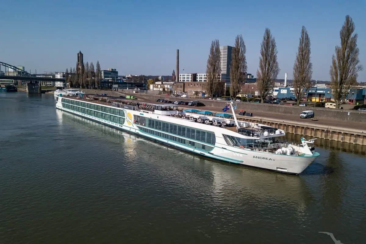 Die MS Andrea, ein weißes Flusskreuzfahrtschiff mit türkisfarbenen Akzenten, schwimmt auf einem breiten Fluss. Im Hintergrund eine Stadtkulisse mit Brücke und Gebäuden unter blauem Himmel.