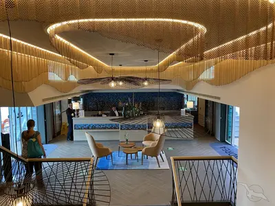 Ein heller, modern gestalteter Empfangsbereich der MS Andrea mit einer Rezeption, Lounge-Sesseln und schwebenden Lichtinstallationen aus goldfarbenen Ketten. Mehrere Personen sind zu sehen.