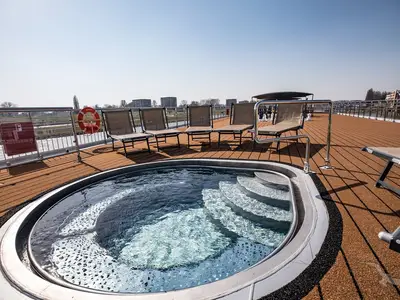 Sonniges Deck der MS Andrea mit rundem Pool samt Treppe und mehreren Liegestühlen. Blick auf den Fluss und die Uferlandschaft bei blauem Himmel.