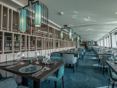 Helles, modernes Restaurant auf der MS Andrea. Viele gedeckte Tische mit blauen Stühlen und Geschirr. Blaue Pendelleuchten und Teppichboden. Große Fenster spenden natürliches Tageslicht.