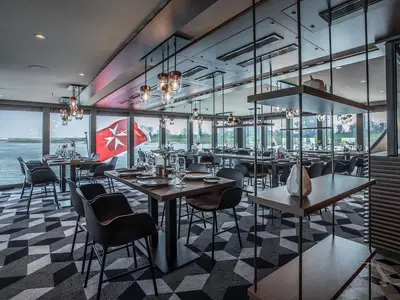 Das helle und moderne Restaurant der MS Andrea bietet mehrere Tische mit Blick auf einen Fluss. Eine rote Flagge mit einem weißen Malteserkreuz ist vor den Fenstern sichtbar.