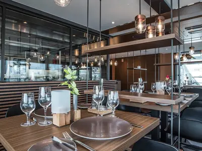 Ein elegantes, helles Restaurant an Bord der MS Andrea mit modernen Holztischen und Stühlen. Zahlreiche Weingläser und gedeckte Teller stehen bereit. Große Fensterfronten bieten Ausblick auf die Umgebung. Eine stilvolle Lichtinstallation hängt von der Decke, ergänzt durch Holzelemente und grüne Pflanzenakzente. Keine Personen sind sichtbar.