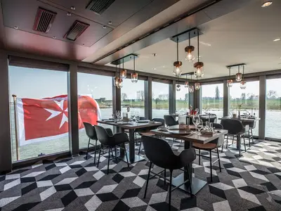Modernes Restaurant an Bord der MS Andrea mit Holztischen und Stühlen. Fensterblick auf Fluss und Ufer. Eine rot-weiße Flagge mit Malteserkreuz ist links sichtbar.