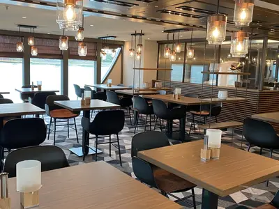 Ein helles und modernes Restaurant an Bord der MS Andrea mit Holztischen, schwarzen Stühlen und großen Fenstern zum Wasser. Stilvolle Kupfer-Hängelampen und ein geometrisches Teppichmuster sind zu sehen.