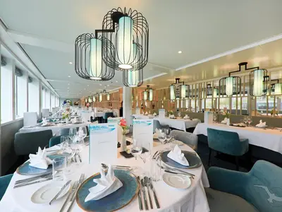 Blick in das elegant gestaltete Restaurant an Bord der MS Andrea. Helle Tische sind mit blauen Tellern und gefalteten Servietten gedeckt. Auffällige Designer-Deckenlampen illuminieren den Raum.
