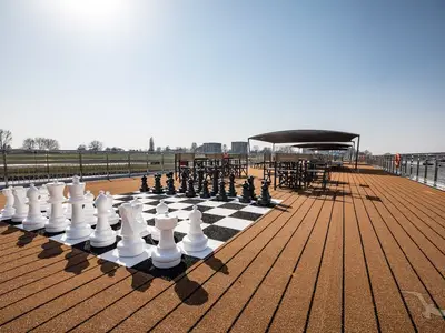 Eine Weitwinkelaufnahme zeigt das Teakholz-Sonnendeck der MS Andrea an einem sonnigen Tag mit einem großen Schachbrett und Sitzgruppen unter Markisen. Das Ufer ist im Hintergrund sichtbar.