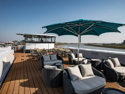 Ein weitläufiges Sonnendeck der MS Andrea mit modernen Rattan-Lounge-Möbeln, Tischen und einem großen türkisen Sonnenschirm. Im Hintergrund fließt ein Fluss vorbei, und man sieht die Steuerbrücke des Schiffs mit einer Person.