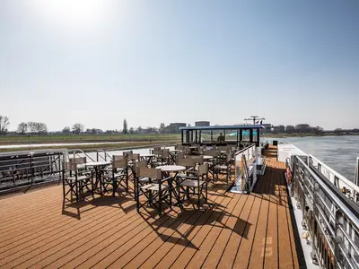 Sonnendeck der MS Andrea mit Tischen und Stühlen auf Holzdeck. Blick auf Fluss, grünes Ufer und entfernte Bebauung unter klarem Himmel. Ein gläserner Aufbau ist sichtbar.