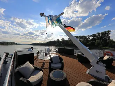 Ansicht vom Sonnendeck der MS Andrea an einem sonnigen Tag. Bequeme Korbsessel stehen auf dem Holzdeck. Im Bildmittelpunkt ein Fahnenmast mit der deutschen Flagge und weiteren bunten Fahnen. Im Hintergrund erstreckt sich ein breiter Fluss, begrenzt von grünen Wäldern unter einem wolkenbedeckten blauen Himmel.