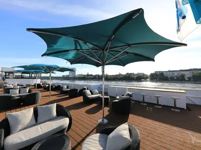 Sonnendeck der MS Andrea mit Lounge-Möbeln, Sonnenschirmen und Bar. Im Hintergrund Fluss und Stadtkulisse. Entspannung an Bord der MS Andrea.
