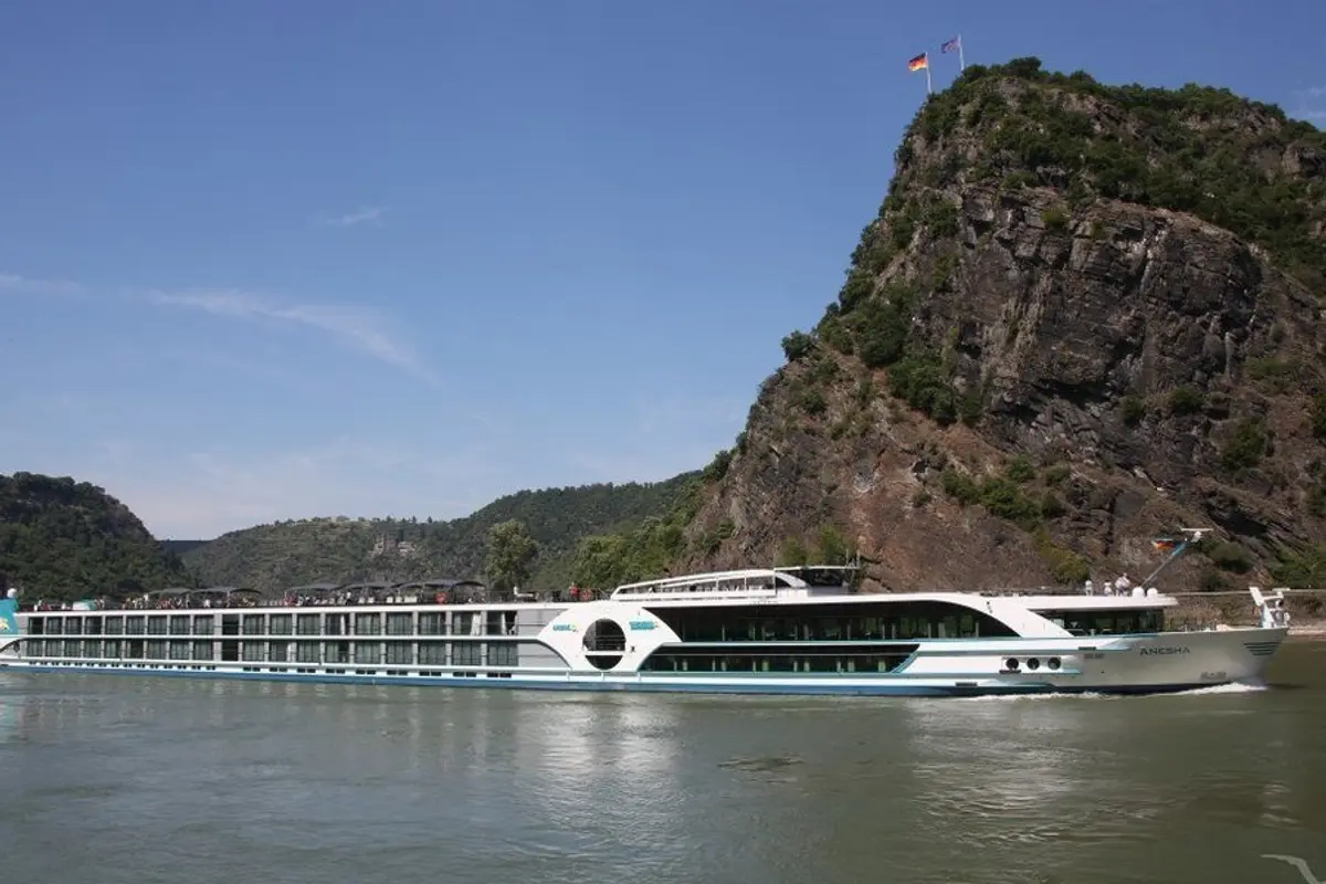 Die MS Anesha, ein modernes Flusskreuzfahrtschiff, fährt auf einem Fluss an bewaldeten Berghängen vorbei. Blauer Himmel und Sonne.