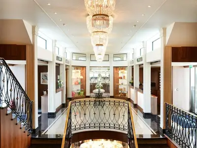 Die luxuriöse Lobby der MS Anesha, von einer Treppe aus. Ein großer Kristallleuchter, flankiert von Geschäften und Rezeptionen, prägt das Bild. Der Marmorboden glänzt.