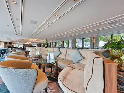 Blick in die stilvolle Panorama-Lounge der MS Anesha mit bequemen cremefarbenen Sofas, Sesseln und runden Glastischen. Große Fenster bieten eine Aussicht auf die Uferlandschaft. Die Decke ist hell mit integrierter Beleuchtung verziert.