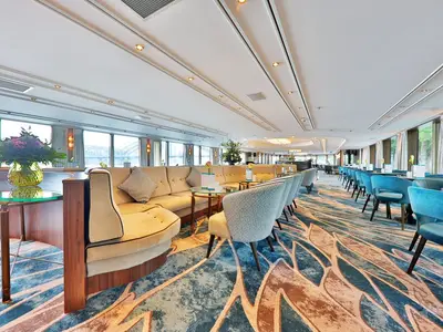 Blick in die helle Panorama-Lounge der MS Anesha mit beigen Rundsofas, türkisfarbenen Stühlen und vielen Fenstern mit Blick nach draußen. Ein Blumenarrangement steht auf einem Glastisch.
