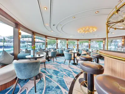Moderne Panorama-Lounge der MS Anesha mit Bar, blauen Sesseln und Tischen. Große Fensterfront bietet Ausblick auf einen Fluss mit Brücke bei Tageslicht.