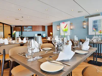 Innenansicht des modernen Restaurants an Bord der MS Anesha mit zahlreichen gedeckten Tischen, eleganten Stühlen, blauer Wand und Kunstwerken.