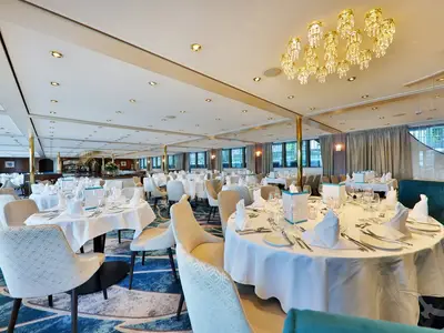Das geräumige 'Vier Jahreszeiten' Restaurant an Bord der MS Anesha zeigt elegant gedeckte Tische, goldene Säulen und eine luxuriöse Einrichtung mit Kronleuchtern. Tageslicht fällt durch die Fenster.