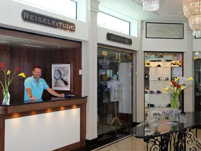 Kreuzfahrtdirektor Mirko Scheffe an der Rezeption der MS Anesha. Er lächelt freundlich und steht hinter dem Tresen mit Blumen und einem Bild. Rechts befindet sich der Eingang zu einem Boutique-Shop.