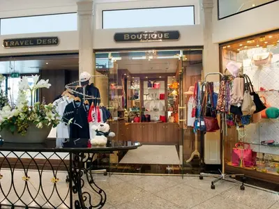 Die helle Boutique an Bord der MS Anesha zeigt Kleidung an Ständern und Accessoires in Vitrinen. Ein Tisch mit Blumen und einem Teddybär ist sichtbar. Schilder "BOUTIQUE" und "TRAVEL DESK" erkennbar.