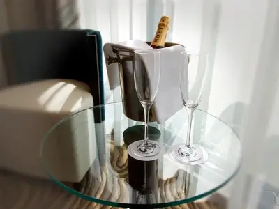 Ein Sektkühler mit einer Flasche Moët und zwei leeren Sektgläsern stehen auf einem runden Glastisch in einer Suite an Bord der MS Anesha. Im Hintergrund sind ein beigefarbener Hocker und ein blauer Stuhl zu sehen, daneben helle Vorhänge.