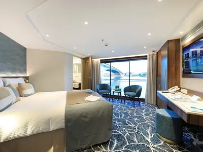 Geräumige Außenkabine der MS Anna Katharina mit großem Fenster zum Fluss. Ein Doppelbett, zwei Sessel, ein Tisch und ein Schreibtisch sind zu sehen.