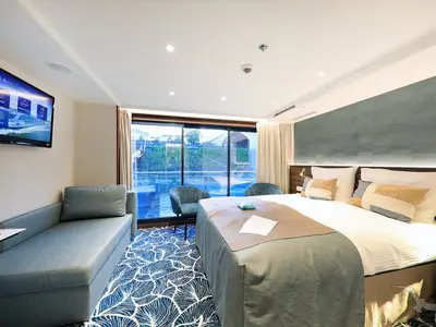Moderne Balkonkabine der MS Anna Katharina mit Doppelbett, Chaiselongue und bodentiefen Fenstern. Blick auf Fluss und Ufer. Einrichtung in Blau und Beige.