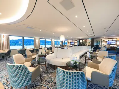 Geräumige und moderne Bar-Lounge an Bord der MS Anna Katharina mit zahlreichen Sitzgelegenheiten und großen Panoramafenstern, die den Blick auf eine bewaldete Flusslandschaft freigeben.
