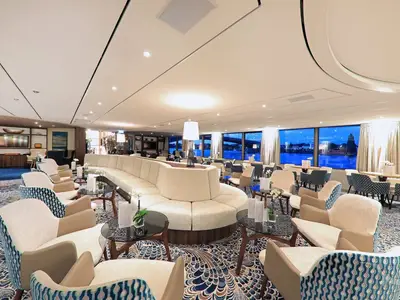 Moderne Bar-Lounge auf der MS Anna Katharina. Gemütliche Sitzgruppen und ein langes Sofa. Große Panoramafenster zeigen eine beleuchtete Flusslandschaft in der Abenddämmerung.