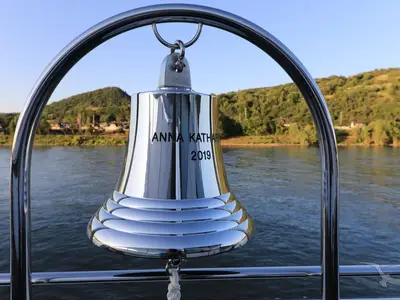 Eine glänzende, silberne Schiffsglocke der MS Anna Katharina mit der Gravur "ANNA KATHARINA 2019" vor dem Hintergrund eines ruhigen Flusses und bewaldeter Hügel. Die Glocke hängt an einem polierten Metallgestell auf dem Schiff.