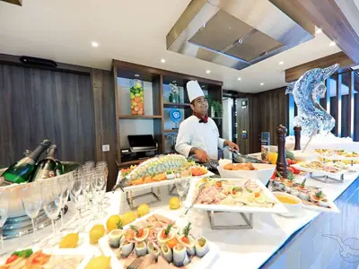 Ein lächelnder Koch bereitet frische Speisen an einem opulenten Buffet auf der MS Anna Katharina zu. Eine Eisskulptur in Delfinform schmückt die Theke, daneben stehen Champagnerflaschen und Gläser bereit.