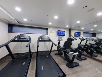 Ein heller Fitnessraum an Bord der MS Anna Katharina mit zwei Laufbändern und mehreren Ergometern, ausgestattet mit Monitoren und einem großen Spiegel an der Wand. Die Geräte stehen auf einem Holzboden.