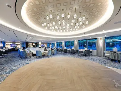 Weitläufige, elegant eingerichtete Lounge der MS Anna Katharina mit heller Holzfläche als Tanzparkett, umgeben von blaugemustertem Teppich. Gemütliche Sessel und Sofas mit Blick durch Panoramafenster ins Grüne.