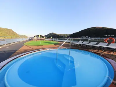 Blick auf das blaue Pool an Bord der MS Anna Katharina auf dem Sonnendeck. Im Hintergrund sind Liegen, ein Mini-Golf-Bereich, der Fluss und grüne Berge mit einem Dorf zu sehen.