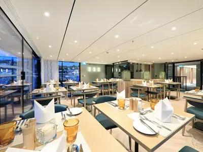 Ein stilvolles Restaurant an Bord der MS Anna Katharina, mit vielen leeren Tischen, großen Panoramafenstern und Blick auf eine beleuchtete Uferpromenade am Abend. Die Tische sind gedeckt mit Besteck, Gläsern und weißen Servietten. Moderne Einrichtung.
