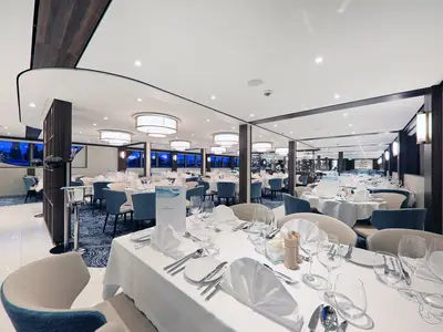 Ein weitläufiges, elegantes Restaurant an Bord der MS Anna Katharina. Zahlreiche Tische sind mit weißen Tischtüchern, Geschirr, Besteck und Gläsern gedeckt. Blaue und cremefarbene Stühle säumen die Tische. Im Hintergrund sind große Fenster zu sehen, die den Blick auf eine abendliche Szene freigeben. Die Decke ist mit modernen, runden Leuchten ausgestattet.