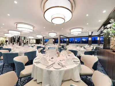 Elegantes Restaurant an Bord der MS Anna Katharina. Weiß gedeckte Tische, blaue Stühle. Moderne Deckenleuchten. Blick durch Fenster auf beleuchtete Skyline bei Nacht.