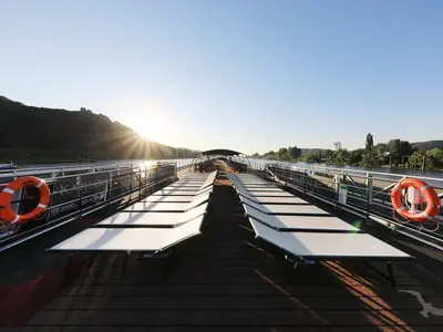 Sonnendeck der MS Anna Katharina bei Sonnenaufgang oder -untergang. Reihen von weißen Liegestühlen bieten Blick auf einen Fluss und bewaldete Hügel. Ein oranger Rettungsring hängt an der Reling.