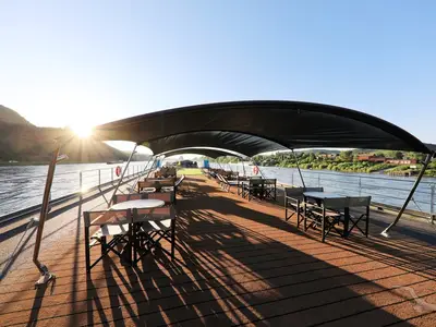Sonnendeck der MS Anna Katharina mit Tischen, Stühlen und schwarzen Sonnensegeln. Lange Schatten auf dem Holzdeck. Fluss und bewaldete Hügel bei Sonnenaufgang.