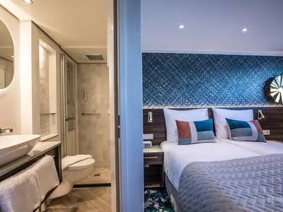 Innenansicht einer modernen Zweibett-Kabine auf der MS Annabelle mit Schlafbereich und Badezimmer. Helle Farben und ein blaues Wanddesign prägen das Ambiente. Das Bad hat eine begehbare Dusche.