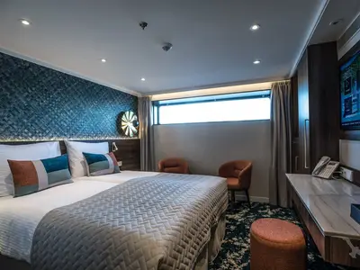 Eine modern eingerichtete Außenkabine an Bord der MS Annabelle. Die Kabine zeigt zwei Einzelbetten mit farbigen Kissen, ein langes Fenster, zwei Sessel und einen Schreibtisch. Die Wand hinter den Betten ist dunkelblau gemustert.