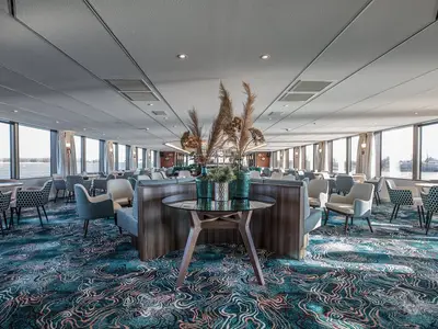 Blick in die helle und moderne Lounge der MS Annabelle mit zahlreichen Sitzgelegenheiten in verschiedenen Blau- und Cremetönen sowie Panoramafenstern zum Fluss.