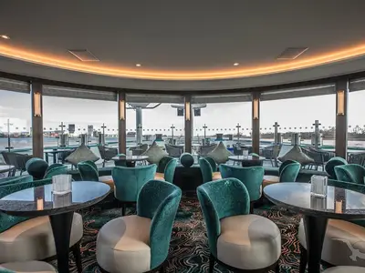 Eine moderne, elegant eingerichtete Bar-Lounge der MS Annabelle mit bequemen türkisfarbenen Sesseln, Marmortischen und großen Panoramafenstern, die den Blick auf das Wasser und eine Hafenumgebung freigeben. Die warm leuchtende Deckenbeleuchtung sorgt für eine gemütliche Atmosphäre.