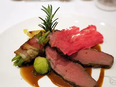 Auf einem weißen Teller liegt ein Hauptgericht: zartrosa Rindfleisch-Tranchen, dazu ein Bund grüner Bohnen im krossen Speckmantel, ein Rosenkohl und ein rosa Rote-Bete-Chip. Eine braune Soße umgibt das Essen an Bord der MS Annabelle.
